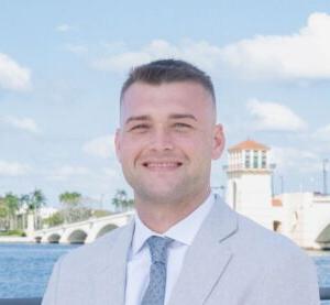 A smiling man in a light gray suit stands by a waterfront with a bridge and palm trees in the background under a blue sky.